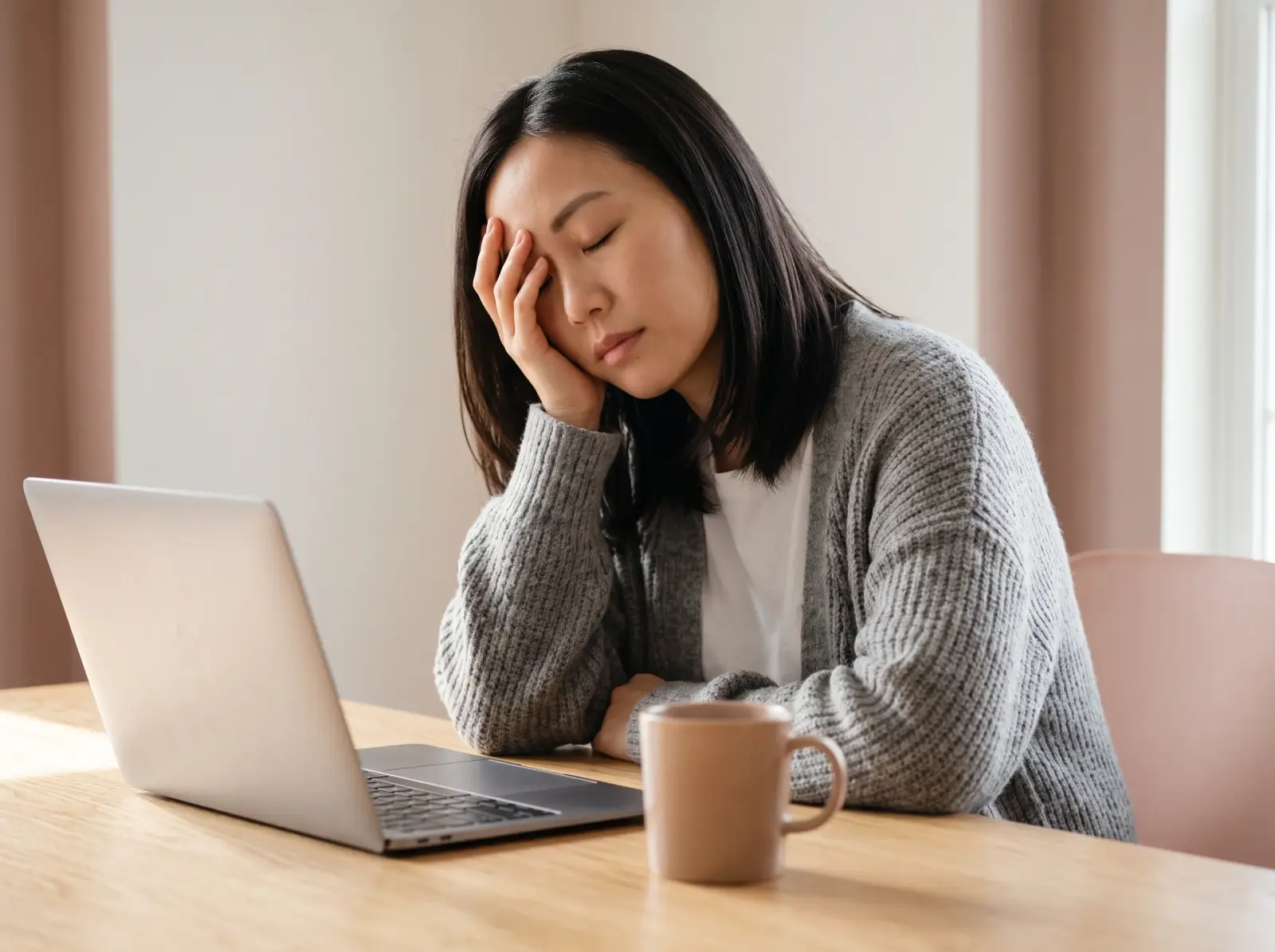 Woman in a grey cardigan resting her head on one hand at a desk beside a laptop