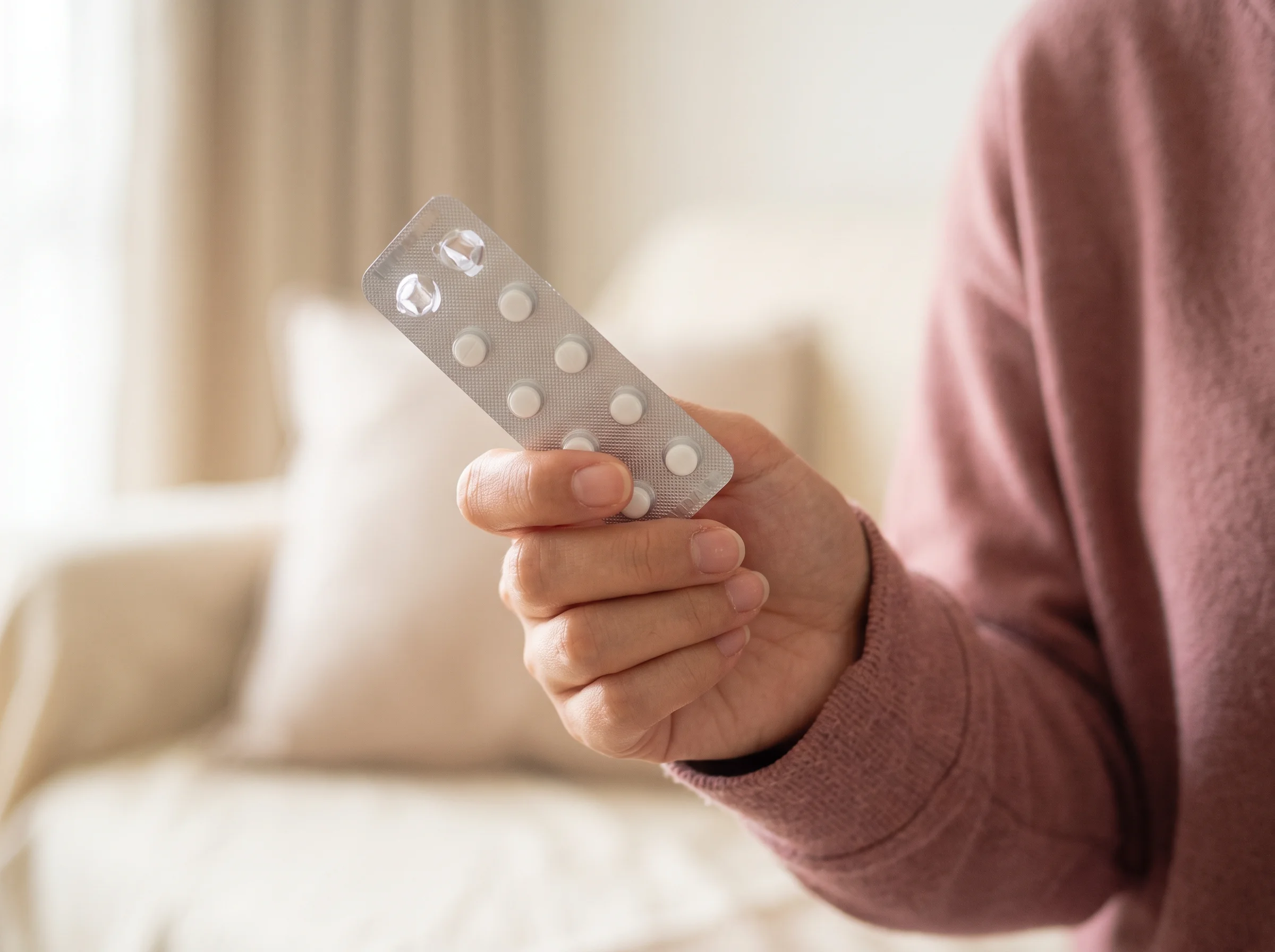 Woman in a pink jumper holding a blister strip of period delay pills on a sofa