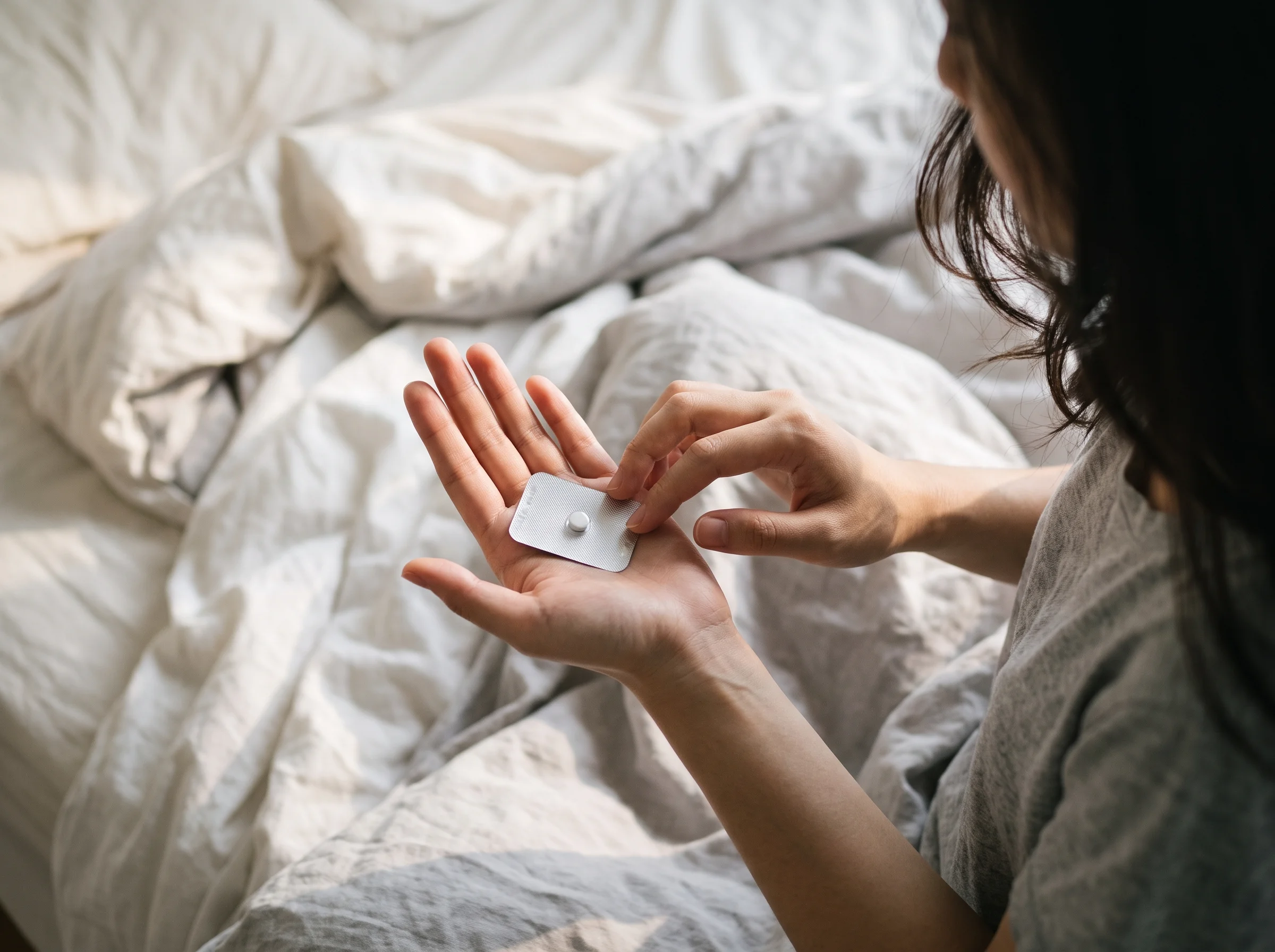Woman sitting on an unmade bed, holding a morning-after pill in her open palm